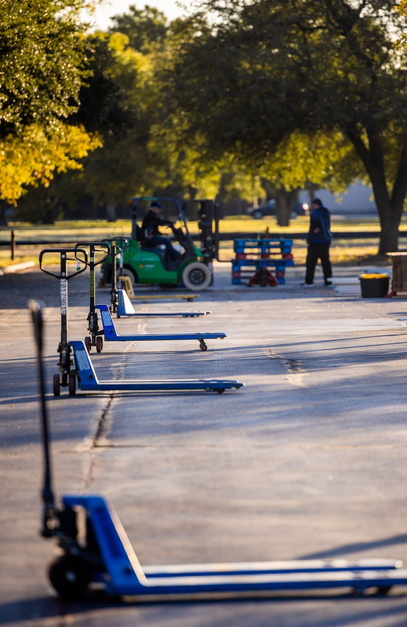 Blue pallet jacks lined up at an outdoor loading area, with a green forklift and workers in the background.