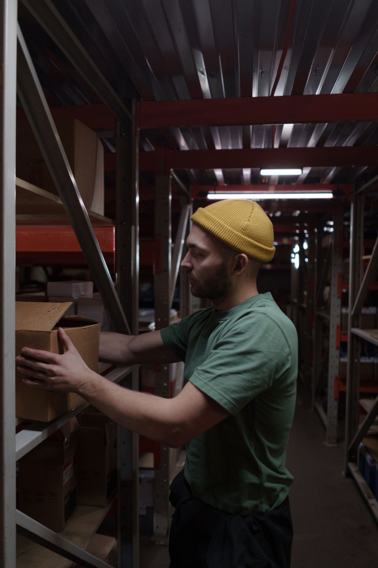 A warehouse worker in a green shirt and yellow knit cap places a carton on a shelf.